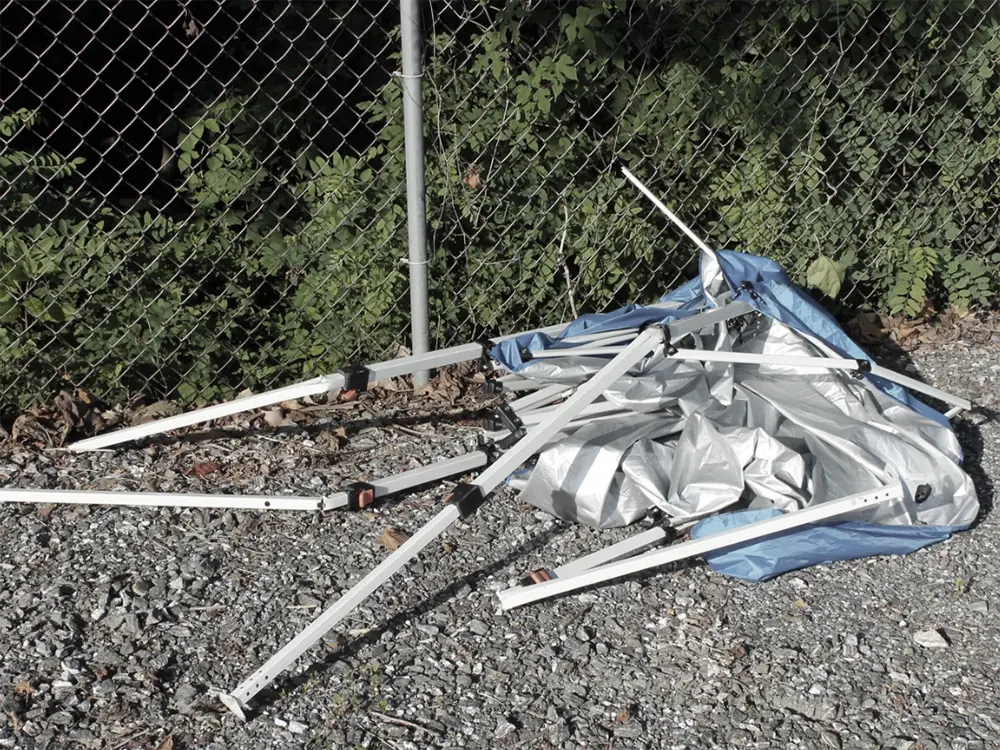 Broken budget gazebo with a snapped leg discarded next to a skip bin
